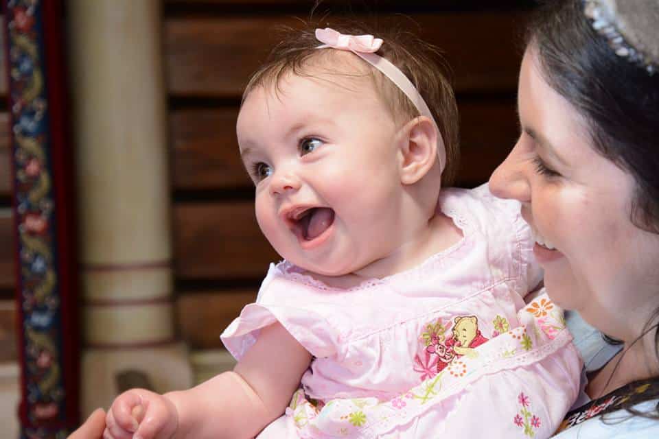 6 month old baby with big smile dressed in pink dress and bow in mom's arms