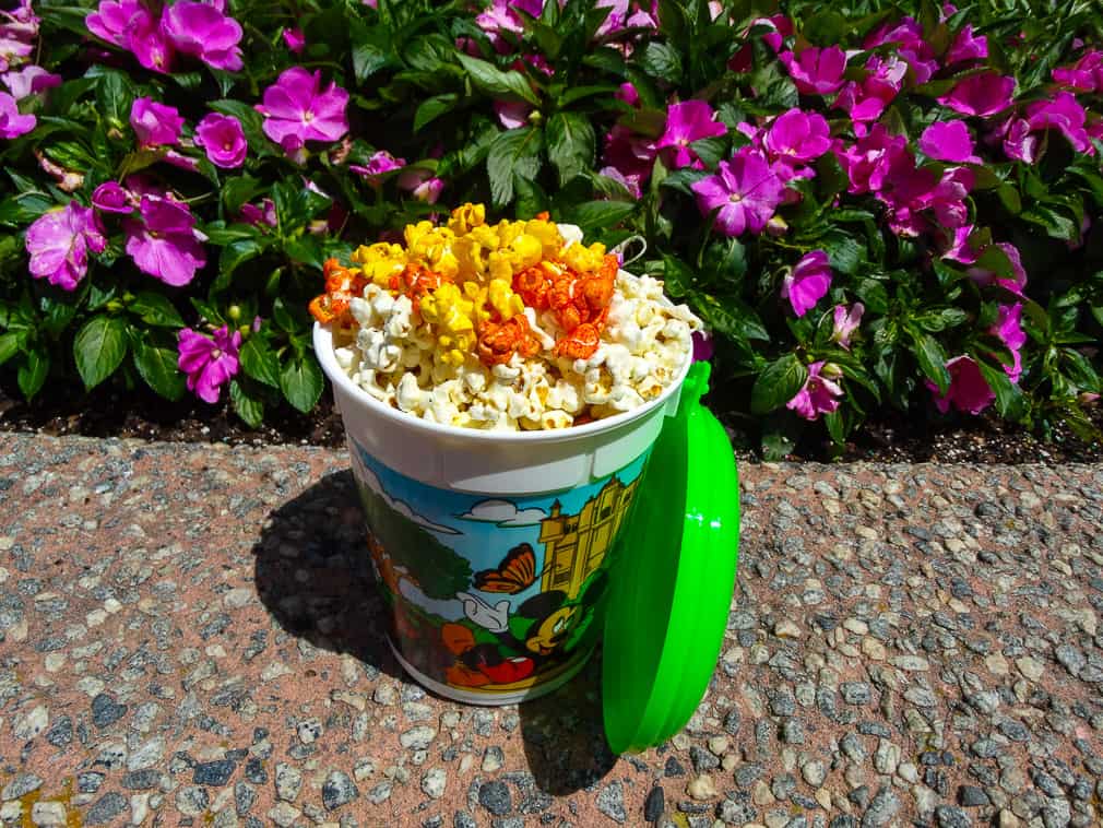 Popcorn bucket with disney characters filled with buffalo, cheddar, and sour cream and chive popcorn with purple flowers in the background