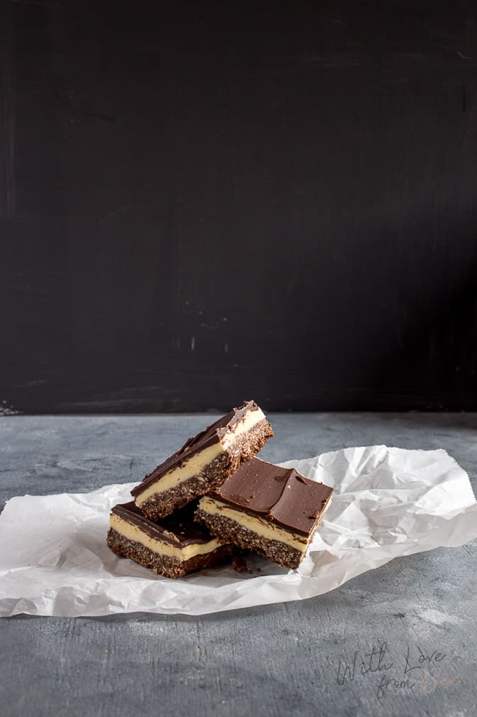 three maple bacon nanaimo bars stacked on top of each other topped with a mint leave against a black and gray background