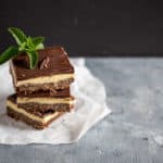 three maple bacon nanaimo bars stacked on top of each other topped with a mint leave against a black and gray background